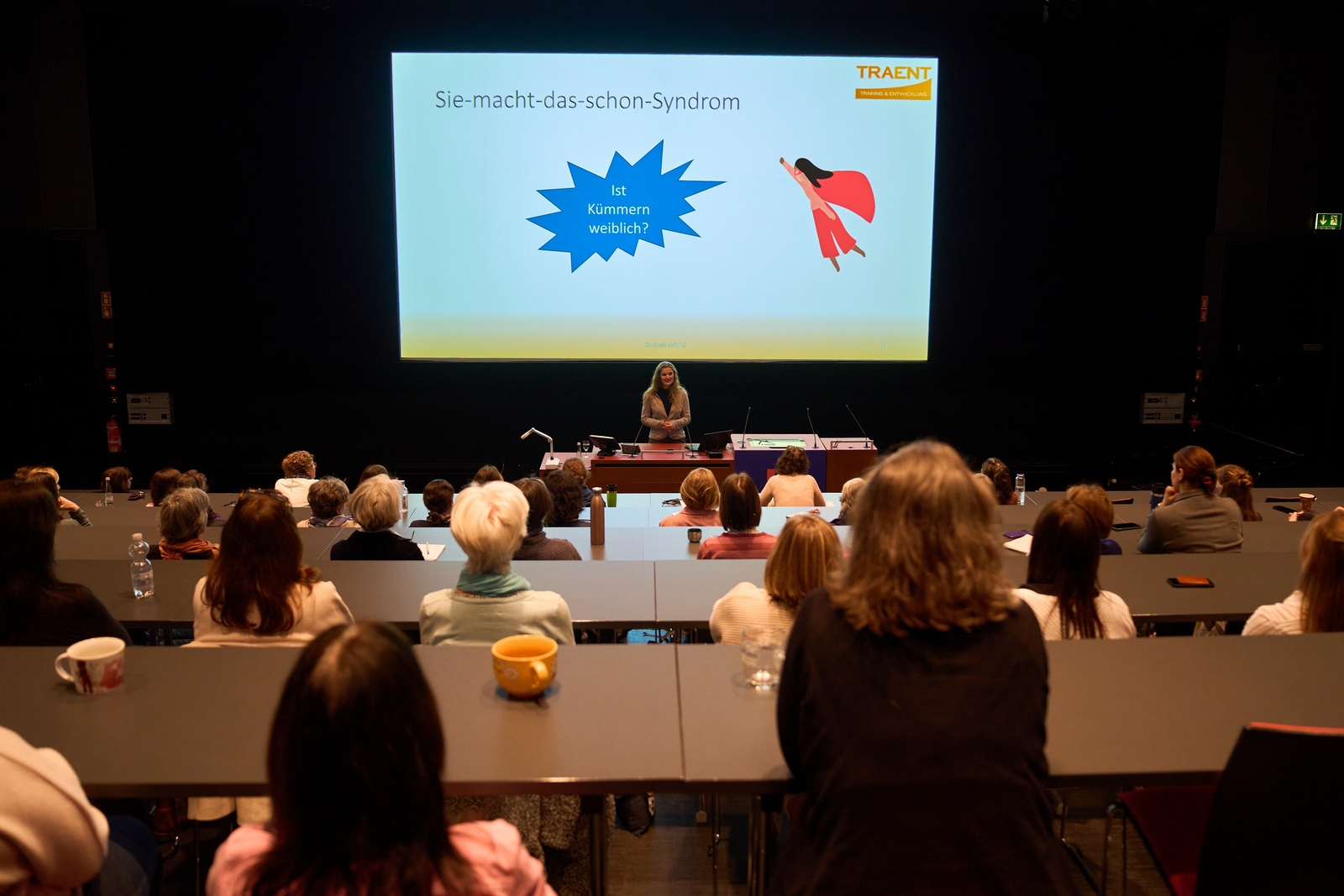 Frauen von hinten sitzen in einem Hörsaal und hören einer vorn unter einer Leinwand mit einer Präsentation stehenden Referentin zu.