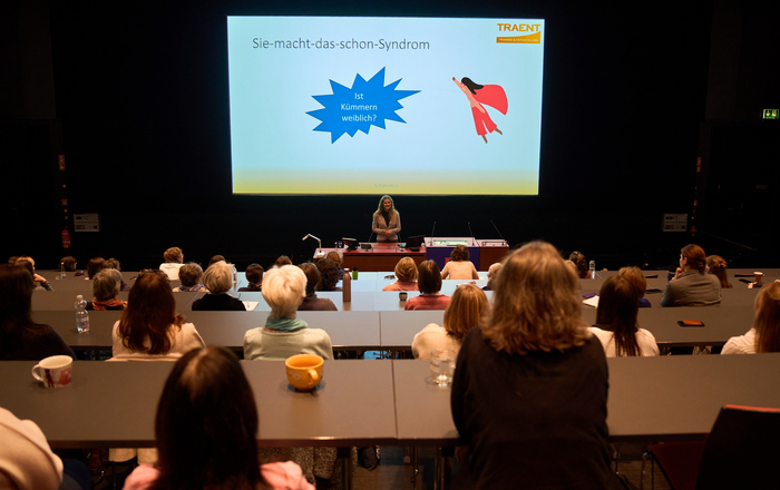 Frauen von hinten sitzen in einem Hörsaal und hören einer vorn unter einer Leinwand mit einer Präsentation stehenden Referentin zu.