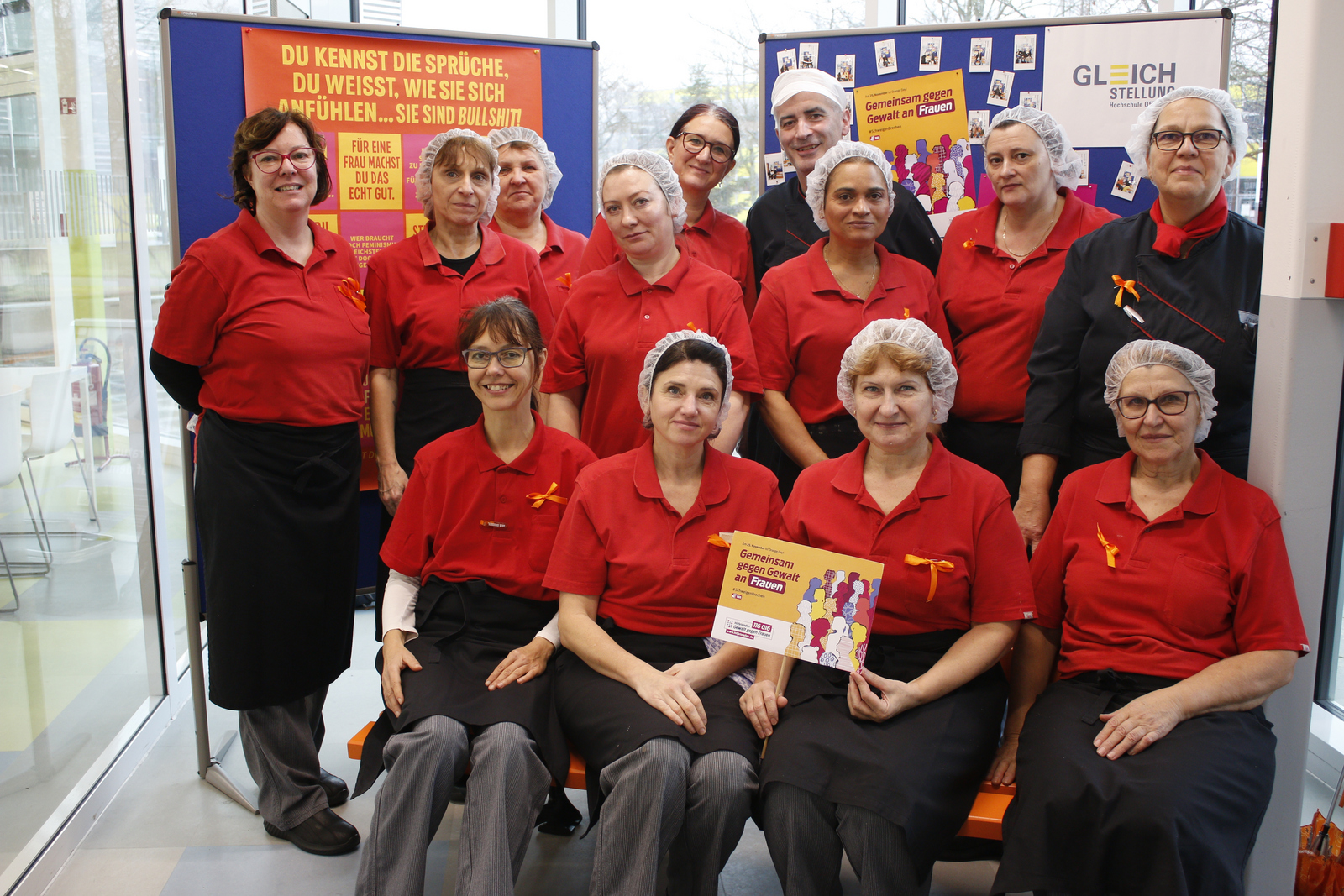 Zwölf Frauen und ein Mann in roten T-Shirts mit orangenen Schleifen stehen hinter und sitzen auf der orangenen Bank 